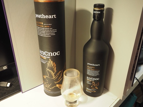 A bottle of AnCnoc Peatheart single malt Scotch Whisky along with its protective and decorative casing, with a small part of the whisky poured into a whisky glass.